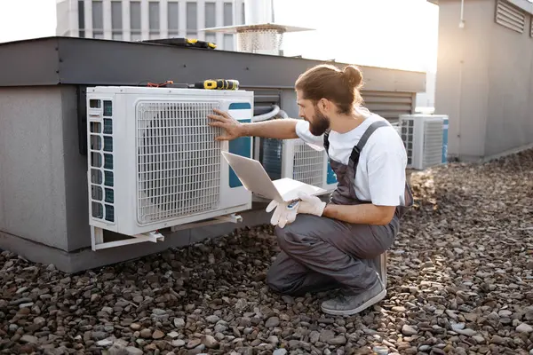 Bearded craftsman in uniform holding digital laptop while checking operation of fan in air conditioner with hand. Caucasian master crouching and using online manual for device outdoors.