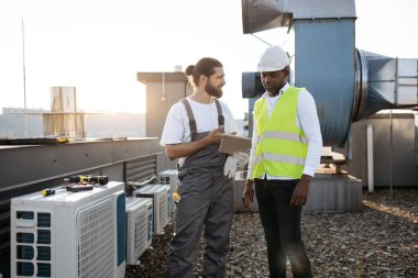 Denetmen bilgilerini gösterirken elinde dijital tablet bulunan usta işçi. Klimalarla çalışan çok kültürlü meslektaşlarım çatıda daha fazla karar alma konusunda hemfikir..