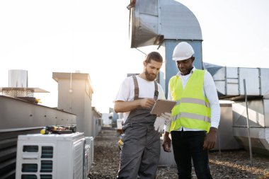 Erkek partnerin yanında duran ve dijital tablette materyaller gösteren deneyimli sakallı bir tamirci. Üniforma giyen çok kültürlü müteahhitler fabrikadaki klimalar hakkında bilgi arıyorlar..