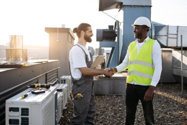 Tulumlu, gülen sakallı bir işçi, elinde modern tabletle siyahi bir mühendisle el sıkışıyor. Çok kültürlü insanlar, tesisin çatısında başarılı bir işbirliği içinde olmaktan memnuniyet duyuyorlar..