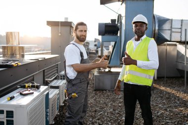 Üniforma giymiş, klimaların yanında duran ve dışarıda dijital tablet kullanırken gülümseyen sanatkarların portresi. Başarılı işbirliği kavramı ve modern araçlar.