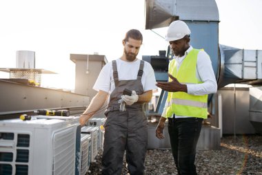 Odaklanmış teknisyen dikilirken ve klima için çoklu sayaç kullanırken erkek denetçi kontrol sürecini kontrol ediyor. Fabrikadaki cihazın voltajını ölçen ve gözlem yapan çok kültürlü tamirciler..