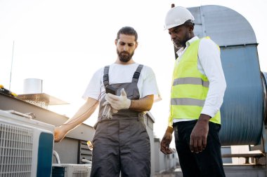 Gri üniformalı ciddi bir zanaatkar, klima için voltaj testi yaparken beyaz şapkalı bir mühendise bakıyor. Çok kültürlü adamlar fabrikada görev yapıyor ve cihazları koruyorlar..