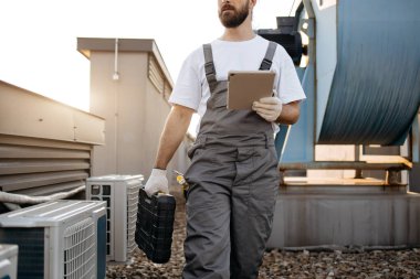Üzerinde fabrika terasında aletler olan siyah bir bavul taşırken elinde kablosuz tablet tutan gri üniformalı sakallı tamirciyi kapatın. Fiziksel çalışma ve modern aygıtlar kavramı.