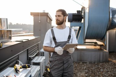 Tulumlu, eldivenli, ayakta duran ve modern tablet kullanan ciddi bir inşaat işçisi. Fabrikanın çatısında aletlerle çalışırken internette sörf yapan beyaz profesyonel bir adam..