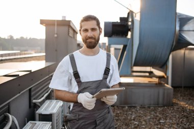 Tulumlu, eldivenli, ayakta duran ve modern tablet kullanan ciddi bir inşaat işçisi. Fabrikanın çatısında aletlerle çalışırken internette sörf yapan beyaz profesyonel bir adam..