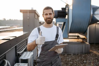Tulumlu, eldivenli, ayakta duran ve modern tablet kullanan ciddi bir inşaat işçisi. Fabrikanın çatısında aletlerle çalışırken internette sörf yapan beyaz profesyonel bir adam..