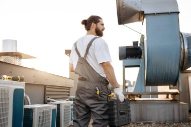 Tulum giymiş tıknaz sakallı efendinin elinde alet çantasını tutarken dikilip baktığı arka manzara. Endüstriyel fabrikanın çatısında odaklanmış erkek denetleme alanı ve bakım cihazları.