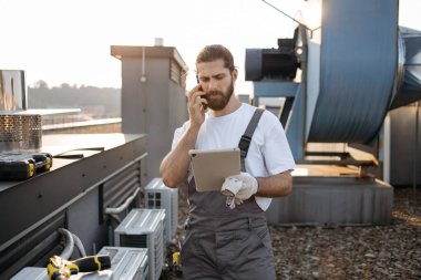 Telsiz cep telefonuyla konuşurken saç örgülü tabletli ciddi bir fabrika işçisi. Beyaz tenli nitelikli adam fabrikanın çatısındaki cihazlarla ilgili sorunları çözüyor.