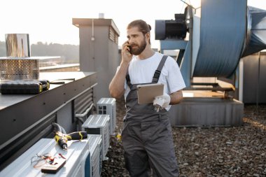 Telsiz cep telefonuyla konuşurken saç örgülü tabletli ciddi bir fabrika işçisi. Beyaz tenli nitelikli adam fabrikanın çatısındaki cihazlarla ilgili sorunları çözüyor.