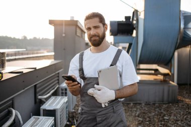 Tulum giymiş sakallı usta ayakta dikilip tablet tutarken aynı zamanda dijital akıllı telefonu temiz havada kullanıyor. Alet kullanan ve fabrikanın çatısında teçhizat taşıyan beyaz yetenekli bir adam..
