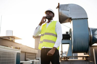 Beyaz kasklı ve yelekli inşaat işçisi ayakta dikilip temiz havada modern akıllı telefona bakıyor. Binanın çatısında fabrika ekipmanlarıyla çalışan zenci profesyonel bir adam..