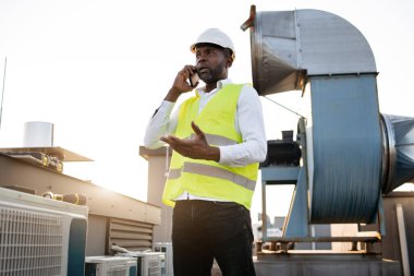 Beyaz miğfer takmış, modern akıllı telefondan sohbet eden Afrikalı Amerikalı ciddi bir mühendis. Yetişkin adam fabrikanın çatısında müşteriyle iş yapıyor ve önemli sorunları çözüyordu..