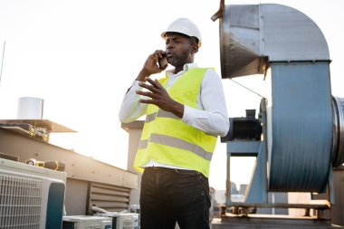 Beyaz miğfer takmış, modern akıllı telefondan sohbet eden Afrikalı Amerikalı ciddi bir mühendis. Yetişkin adam fabrikanın çatısında müşteriyle iş yapıyor ve önemli sorunları çözüyordu..