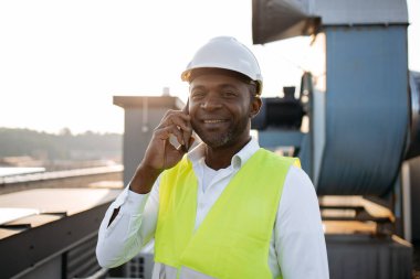 Temiz havada dijital cep telefonunda konuşurken ayakta durup gülümseyen iş üniforması giymiş erkek işçinin portresi. Afrikalı Amerikalı adam konuşuyor ve fabrikanın çatısında cihazlarla çalışıyor..