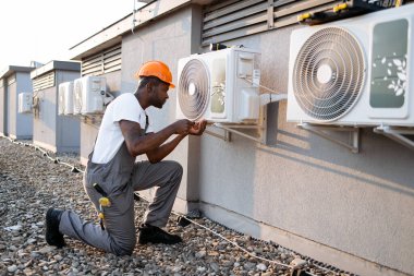 Vasıflı müteahhit havalandırmayı denetliyor ve tamirat sırasında tornavida kullanıyor. Siyahi ciddi bir erkek tek dizinin üstünde duruyor ve binanın çatısında tamir cihazını tamir ediyor..