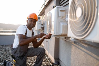 Afrika kökenli Amerikalı uzman bir tamirci, tek dizinin üstünde duruyor ve elinde tornavidayla binanın çatısında klima tamir ediyor. Fabrika bakımı kavramı.