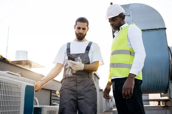 Gri üniformalı ciddi bir zanaatkar, klima için voltaj testi yaparken beyaz şapkalı bir mühendise bakıyor. Çok kültürlü adamlar fabrikada görev yapıyor ve cihazları koruyorlar..
