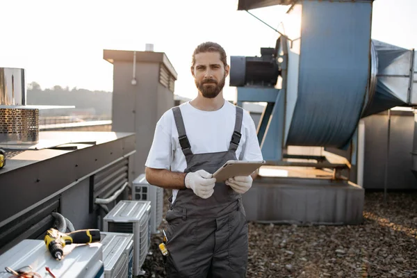 Tulumlu, eldivenli, ayakta duran ve modern tablet kullanan ciddi bir inşaat işçisi. Fabrikanın çatısında aletlerle çalışırken internette sörf yapan beyaz profesyonel bir adam..