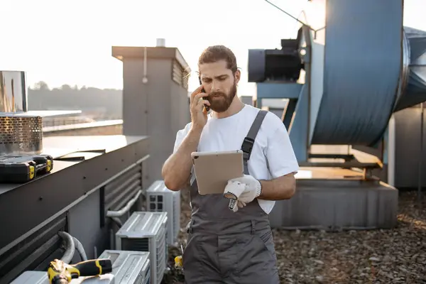 Telsiz cep telefonuyla konuşurken saç örgülü tabletli ciddi bir fabrika işçisi. Beyaz tenli nitelikli adam fabrikanın çatısındaki cihazlarla ilgili sorunları çözüyor.