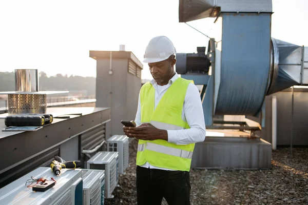 Busy construction worker in white helmet and vest standing and looking at modern smartphone on fresh air. Black professional man working with factory equipment at industry in roof of building.