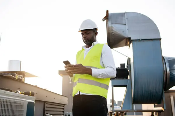Busy construction worker in white helmet and vest standing and looking at modern smartphone on fresh air. Black professional man working with factory equipment at industry in roof of building.