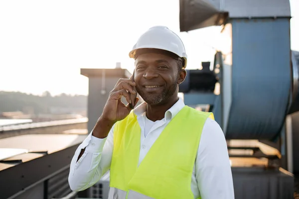 男工穿着工作服站着笑着在数字移动电话上呼吸新鲜空气的画像 非洲裔美国人与工厂屋顶上的设备进行对话和工作 — 图库照片