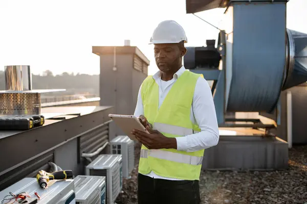 Competent craftsman wearing white hard hat standing and holding wireless tablet in hands outdoors. Black concentrated man searching for information in device and monitoring work at plant in roof.