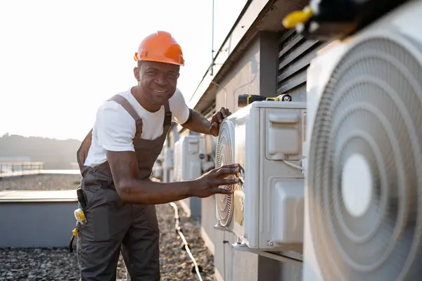 African american factory worker in orange hard hat standing and holding hands on air conditioner while checking attachment to wall outdoors. Concept of manual work and installation of equipment.