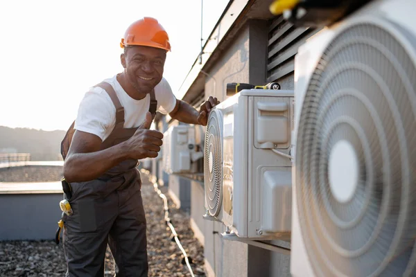 African american factory worker in orange hard hat standing and holding hands on air conditioner while checking attachment to wall outdoors. Concept of manual work and installation of equipment.