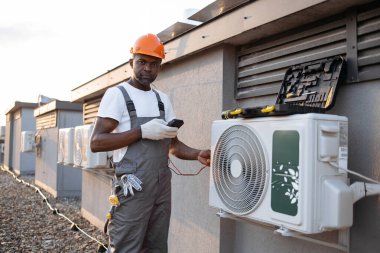 Gri tulumlu inşaat mühendisi elinde dijital çoklu metre tutuyor ve açık havada klima dağıtırken voltaj seviyesini kontrol ediyor. Afrika kökenli Amerikalı tamirci çatıya modern cihazlar yerleştiriyor..