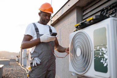 Konut binasının çatısına modern soğutma sistemi kurarken voltaj testini kullanan odaklanmış bir Afro-Amerikan uzmanı. Asılan klimada mevcut gücü ölçen yönetici.