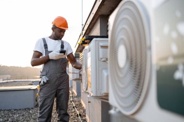 Konut binasının çatısına modern soğutma sistemi kurarken voltaj testini kullanan odaklanmış bir Afro-Amerikan uzmanı. Asılan klimada mevcut gücü ölçen yönetici.