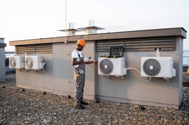 Üzerinde alet çantası olan beyaz klimanın yanında duran meşgul ustabaşının tam boy görüntüsü. Yetenekli Afro-Amerikan erkek fabrika ekipmanlarının gücünü test etmek için dijital çokölçer kullanıyor..