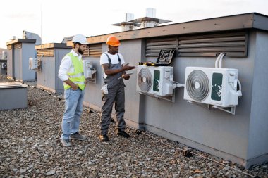 Elinde tabletle Afrika kökenli Amerikalı teknisyen çatıdaki beyaz şapkalı Kafkasyalı yöneticiye tamir edilmiş klimayı gösteriyor. Fabrika işçileri modern ekipmanları korumak için işbirliği yapıyorlar..