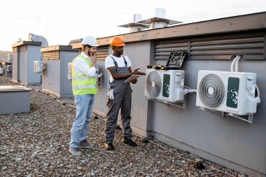 Elinde tabletle Afrika kökenli Amerikalı teknisyen çatıdaki beyaz şapkalı Kafkasyalı yöneticiye tamir edilmiş klimayı gösteriyor. Fabrika işçileri modern ekipmanları korumak için işbirliği yapıyorlar..
