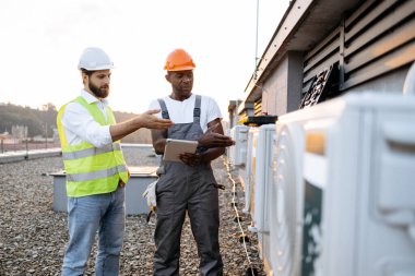 Çok kültürlü erkekler, açık havada bozuk soğutma sisteminin yanında dururken birbirlerine klima tamiri hakkında tavsiyelerde bulunuyorlar. Kullanıcı bilgili web sitelerini kaydırmak için tablet kullanan adamlar.