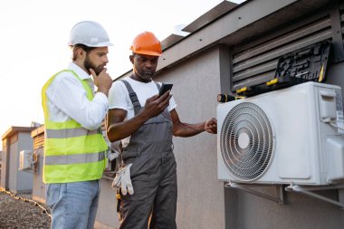 İlgilenen meslektaşım dışarıda modern cihazlara bakarken dikkatli bir işçi dijital çokölçer tutuyor. Çok ırklı tamirciler çatıdaki yeni klimayı tamir ediyor..