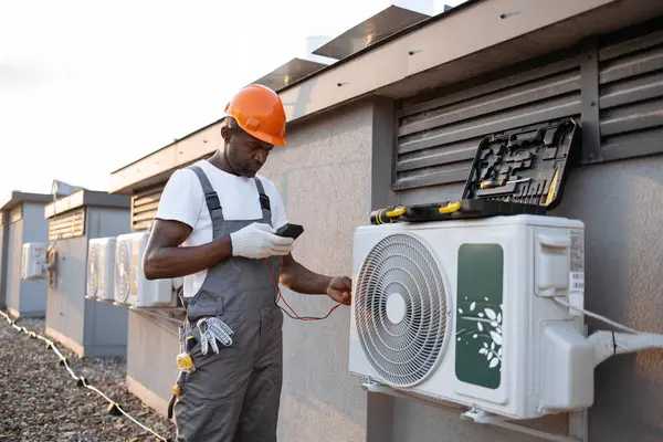 Busy civil engineer dressed in gray jumpsuit holding digital multimeter and checking voltage level in handing air conditioner outdoors. African american repairman installing modern device on rooftop.