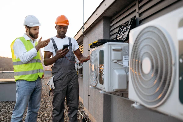 İki çok ırklı teknisyenden oluşan ekip voltajı modern multimetre ile kontrol ediyor ve soğutma sisteminin onarımını tartışıyorlar. Alet çantalı becerikli erkek müfettişler dışarıdaki klimaya bakıyor..