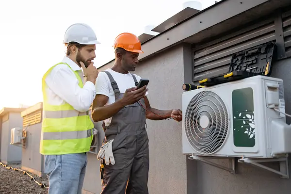 İlgilenen meslektaşım dışarıda modern cihazlara bakarken dikkatli bir işçi dijital çokölçer tutuyor. Çok ırklı tamirciler çatıdaki yeni klimayı tamir ediyor..