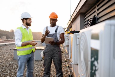 Koruyucu şapkalı çok ırklı inşaat yöneticileri, klima tamiri hakkında modern tabletten bilgi okuyorlar. Sanayi fabrikasının çatısında duran iki adam..