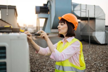 Turuncu kask takan kadın ve sarı güvenlik yeleği giyen, geniş soğutma sisteminde tornavida takan kadın görüntüsü. Beyaz kadın klima tamir etmek için yetenekler kullanıyor..