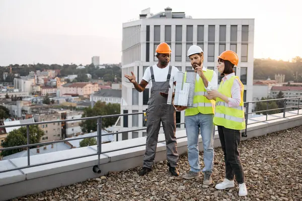 Stress-resistant three construction workers wearing reflective vests looking at small model of residential building outdoors. Multiracial team of determined managers standing on rooftop.