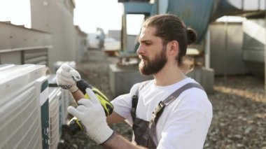 Sakallı bir zanaatkar fabrikanın çatısındaki klimayı tamir ederken eğilip matkap kullanıyor. Üniformalı beyaz erkek, makine ekipmanlarını tamir ediyor ve geliştiriyor..