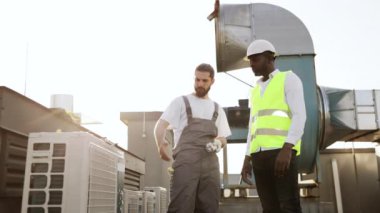 Beyaz miğferli ve yansıtıcı yelekli Afro-Amerikan mühendis Kafkasyalı tamircinin çalışmalarını değerlendirirken klimaya bakıyor. Bitki ekipmanları ve bakım konsepti.