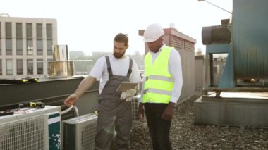 Denetmen bilgilerini gösterirken elinde dijital tablet bulunan usta işçi. Klimalarla çalışan çok kültürlü meslektaşlarım çatıda daha fazla karar alma konusunda hemfikir..