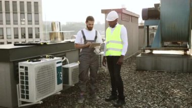 Erkek partnerin yanında duran ve dijital tablette materyaller gösteren deneyimli sakallı bir tamirci. Üniforma giyen çok kültürlü müteahhitler fabrikadaki klimalar hakkında bilgi arıyorlar..