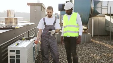 Odaklanmış teknisyen dikilirken ve klima için çoklu sayaç kullanırken erkek denetçi kontrol sürecini kontrol ediyor. Fabrikadaki cihazın voltajını ölçen ve gözlem yapan çok kültürlü tamirciler..