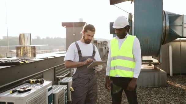 アフリカ系アメリカ人のスーパーバイザーと お互いの隣に立って 現代のタブレットの画面を見ています 屋上にエアコンについての情報を読む修飾された工場労働者 — ストック動画